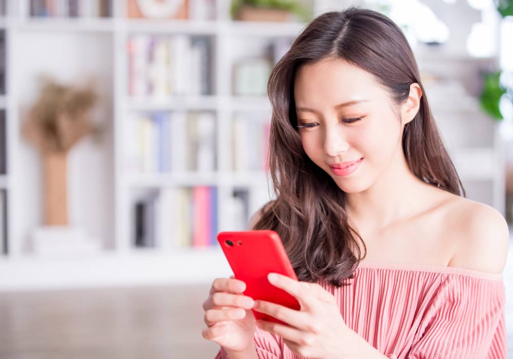 部屋の中でスマートフォンを操作しながら、笑顔でオンラインのドレスレンタルサービスを利用している女性の様子