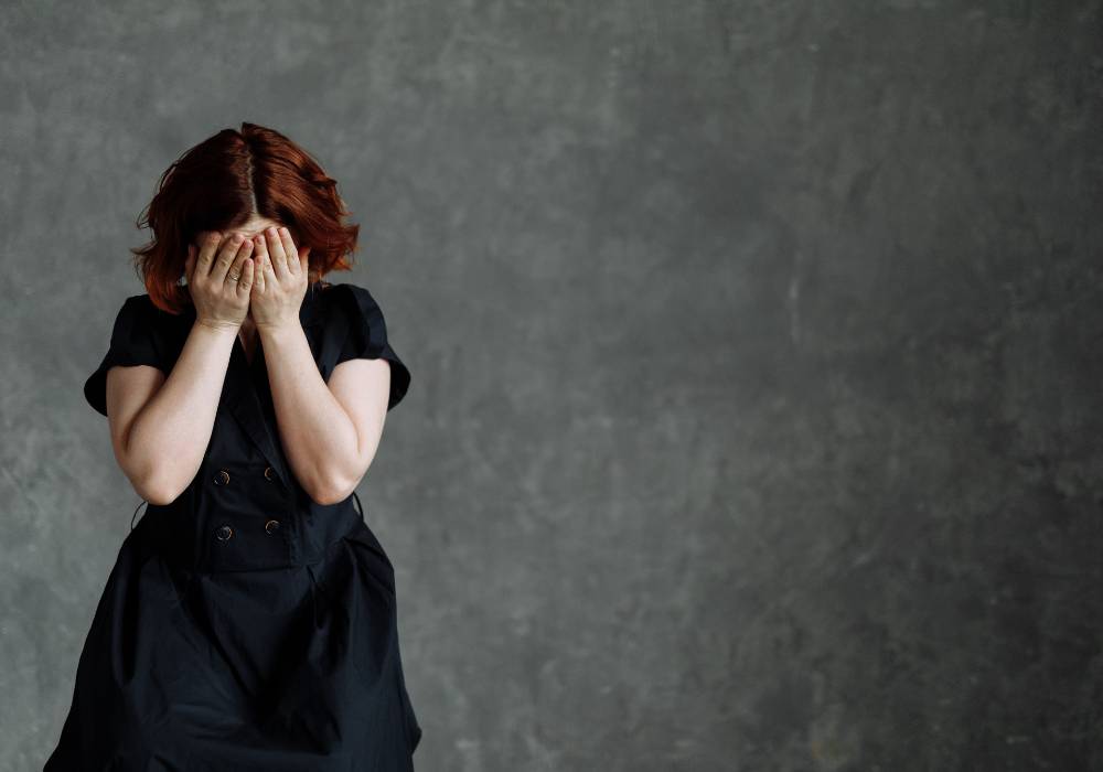 グレーの壁を背景に、黒いワンピースを着て両手で顔を覆いながら悩んでいるような女性の姿