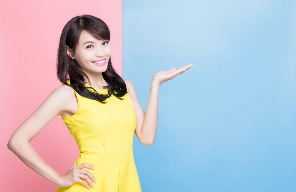 ピンクとブルーの2色背景の前で、鮮やかなイエローのドレスを着て、自信に満ちた笑顔で何かを紹介するポーズをとる20代の女性