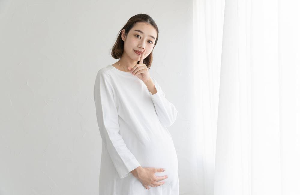 人差し指を口元に当てて微笑む、白いワンピースを着た妊婦の女性