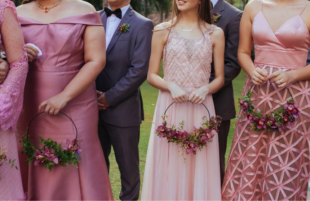 結婚式やパーティーの参列者たちが並んで立っている。ピンク色の様々なデザインのドレス（オフショルダー、刺繍入り、幾何学模様）を着用した女性たちと、グレーのスーツを着用した男性が、花輪のハンドブーケを持っている。